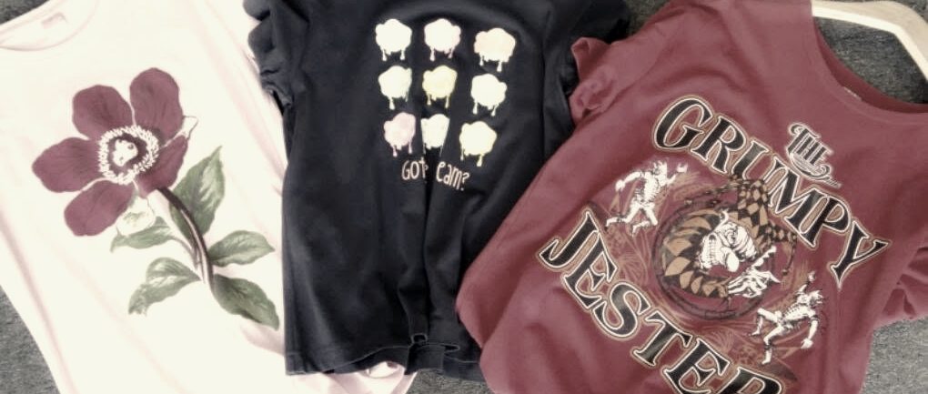 Three printed t-shirts on hangers: a pink flower, pastel icons on navy and a red jester design