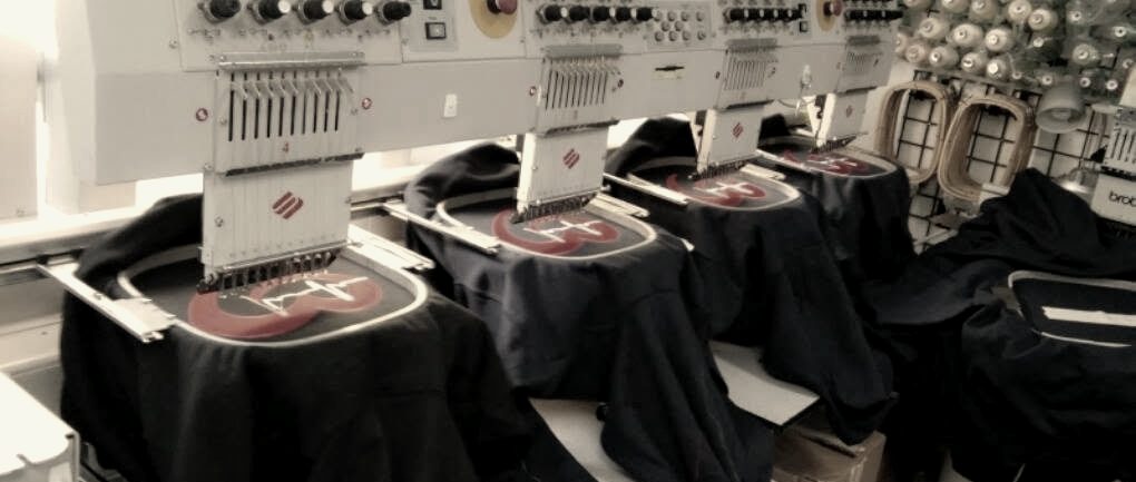Navy jackets hooped across a multi-head embroidery machine, red logos stitching, thread wall behind