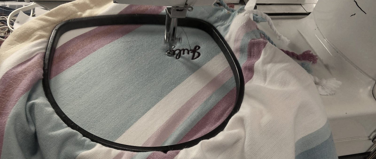 A striped pink and aqua beach towel hooped on an embroidery machine, a small script name stitched