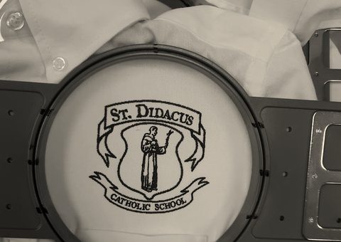 A school crest stitched in maroon on a white shirt, still in the hoop
