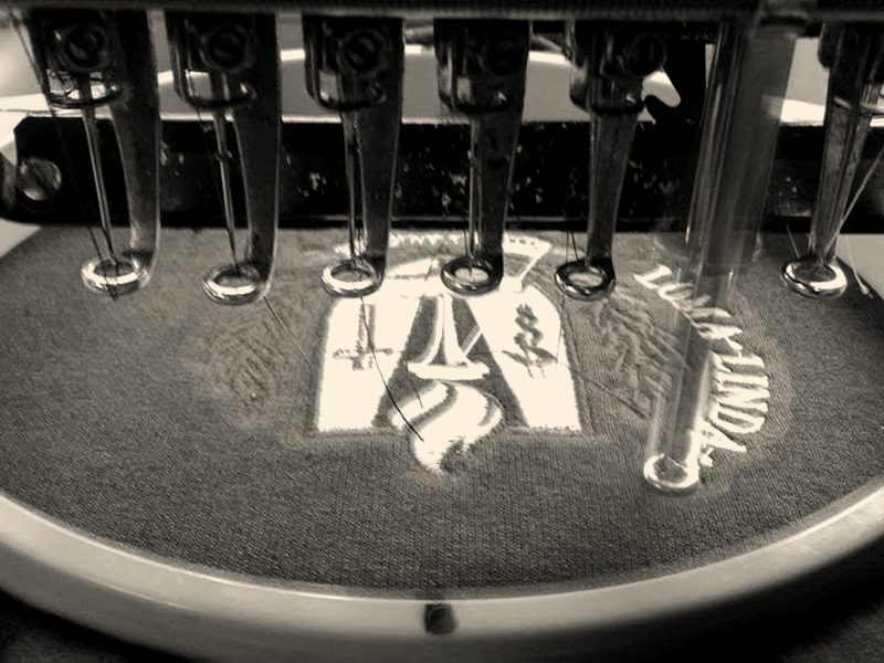 Needles stitching a red and white crest on navy fabric in a round hoop