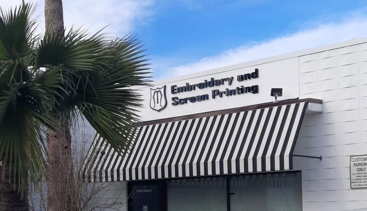 The MMM Embroidery storefront in Newhall Square: a white building with an 'Embroidery and Screen Printing' sign, the shield logo and a black-and-white striped awning beside a palm tree